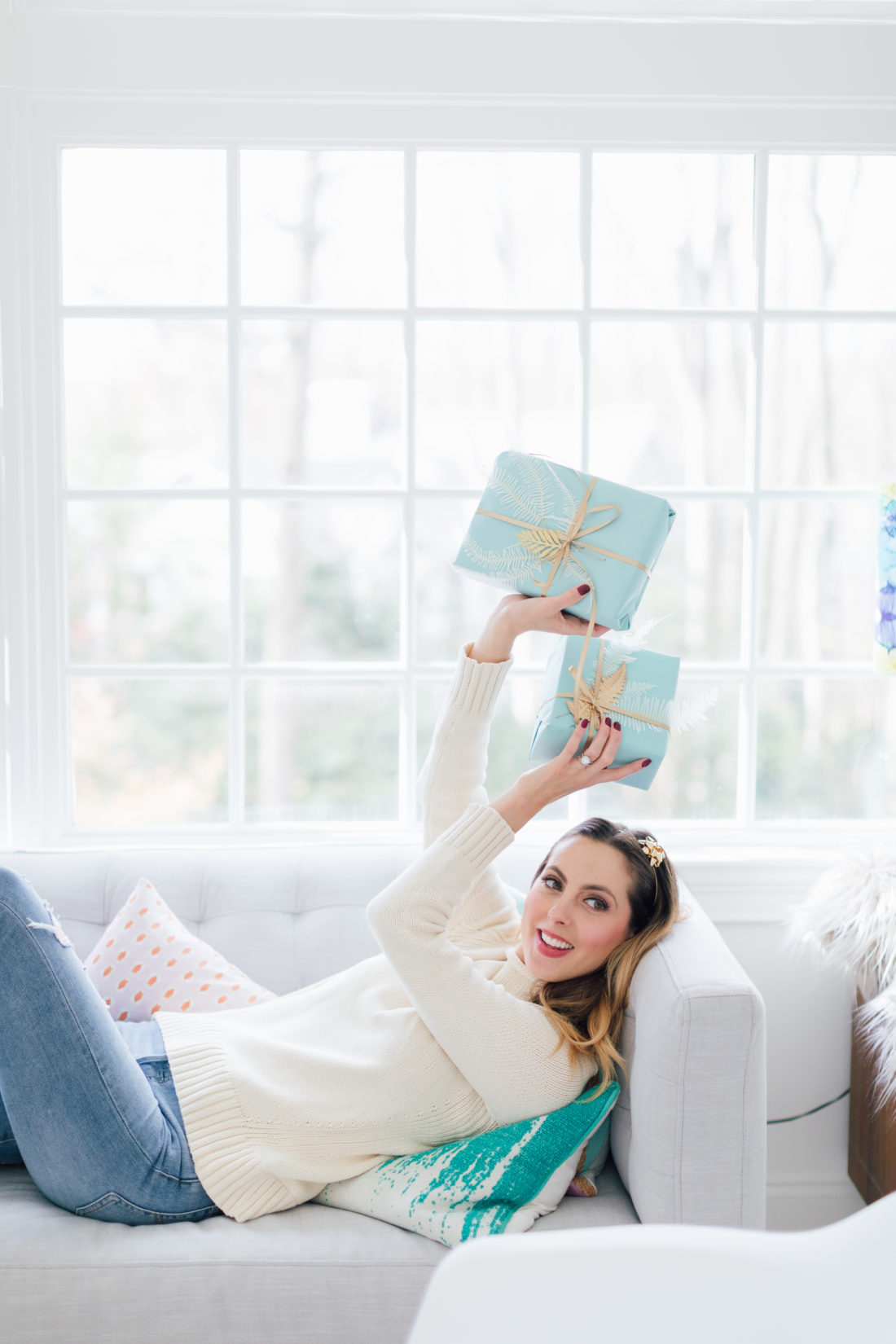 Eva Amurri Martino wears her cream colored sweater and levi jeans, and lays on the couch at home, holding robin's egg colored gifts with gold ribbon