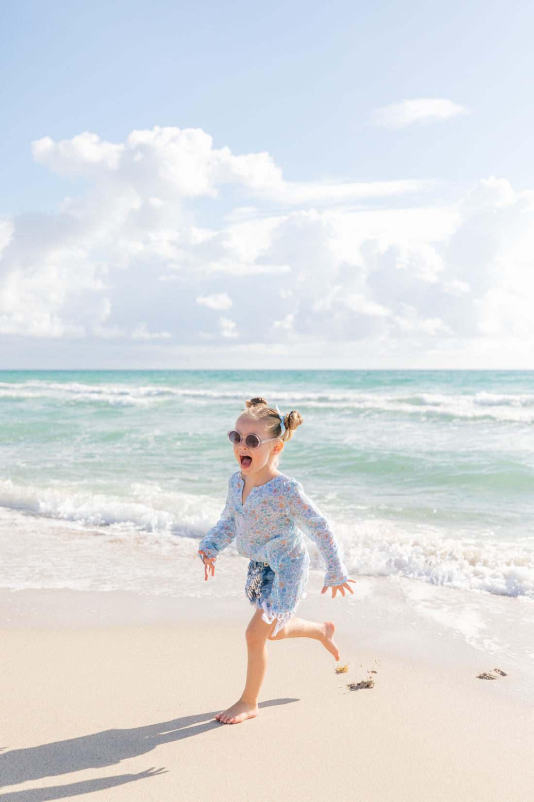 Marlowe Martino wears a blue, floral tunic from the Happily Eva After x Masala Baby capsule collection, and denim shorts, and frolics in the sand and surf beachside in Miami